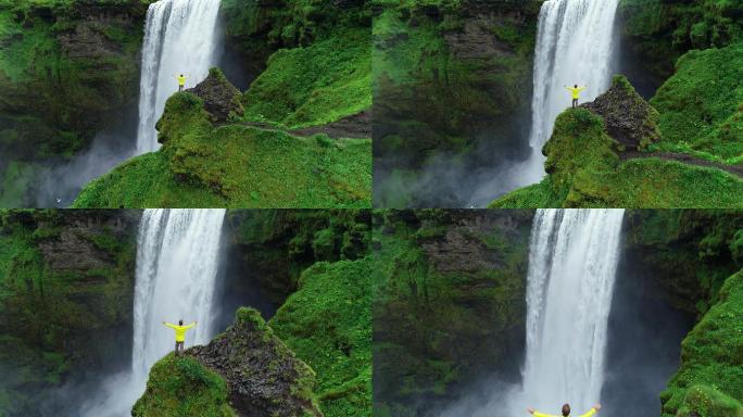 一名男子举起双臂站在瀑布前的悬崖顶上