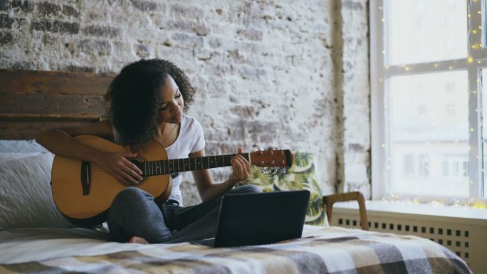 少女坐在家里的床上，用笔记本电脑学习吉他