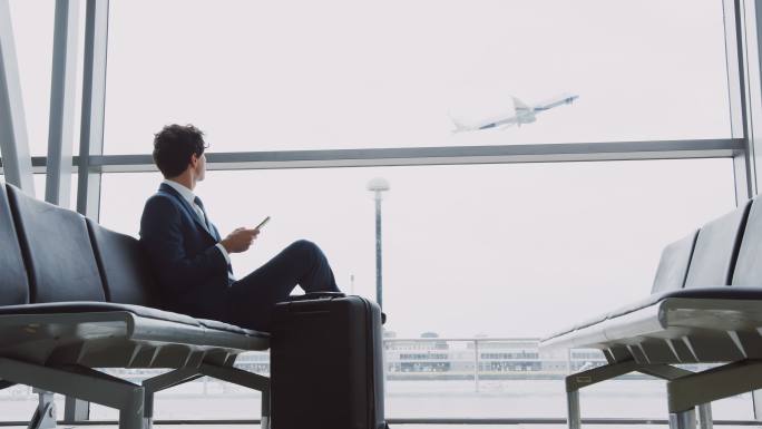 商人坐在机场候机厅使用手机，背景飞机起飞