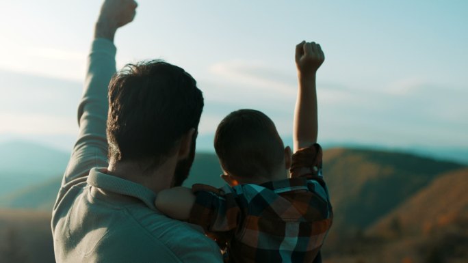 父亲抱着儿子，他们在山顶上举起双手