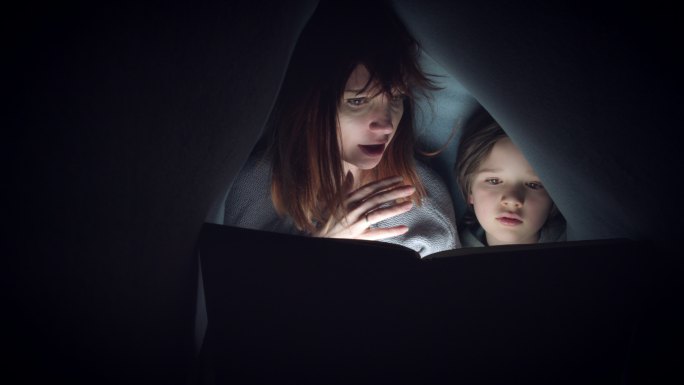 孩子和妈妈在毛毯下读恐怖故事书
