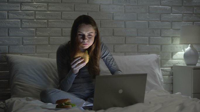 年轻女性晚上在床上上网时吃汉堡包