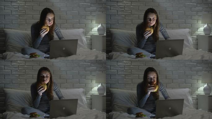 年轻女性晚上在床上上网时吃汉堡包