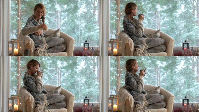 年轻的女人喝着咖啡穿着毛衣坐在大窗前