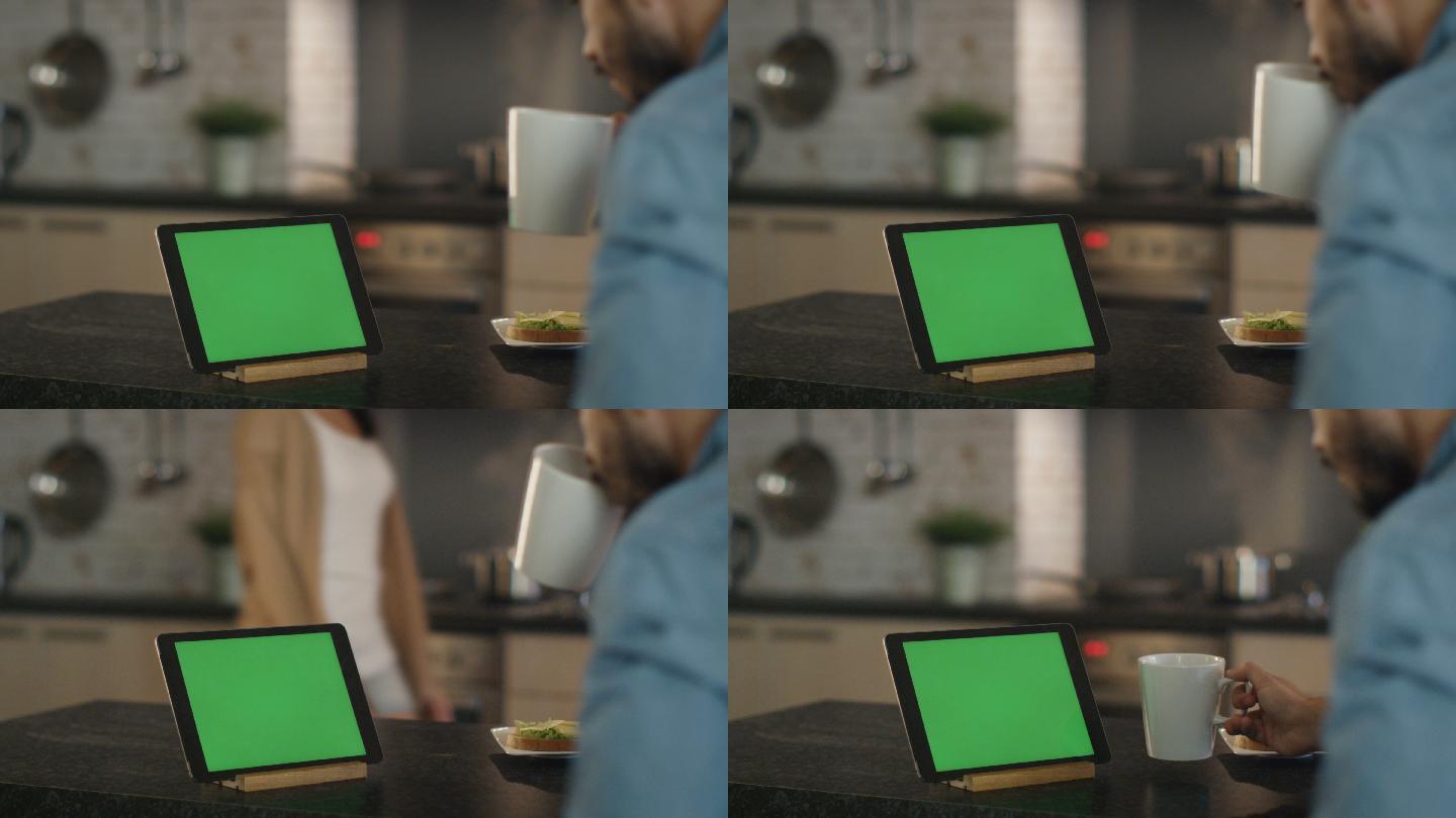 年轻人坐在绿色屏幕的平板电脑前啜饮着杯子