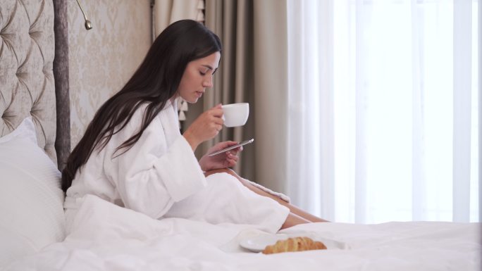 一个年轻的女人坐在床上看电话，喝咖啡
