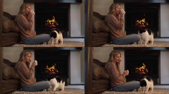 女子坐在壁炉边喝咖啡，小猫正在蹭她的腿