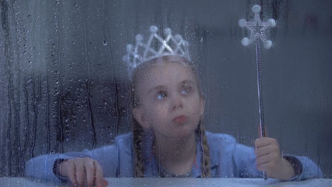 拿着魔杖的小女孩独自坐在雨天的窗户后面