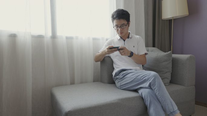 4K年轻男性在窗户旁边沙发上玩手机游戏