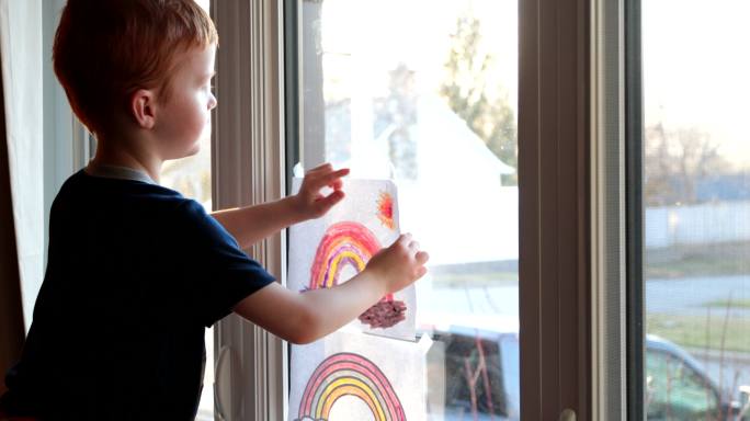 一个小男孩把他的彩虹画贴在家里的窗户上