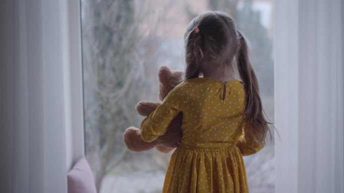 小女孩抱着泰迪熊望着窗外的后视图。