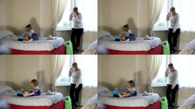 小女孩在床上玩玩具，妈妈站在窗口打电话