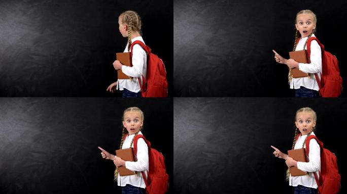 震惊的小女孩背着背包拿着书，手指着黑板