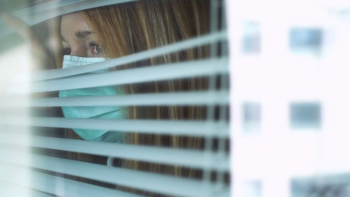 悲伤的年轻女子戴着医疗口罩看着窗外