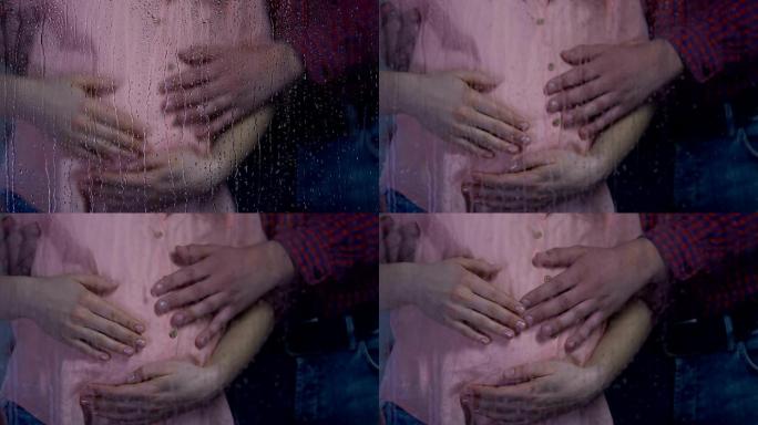 怀孕的女人和男人在雨窗后抚摸着肚子