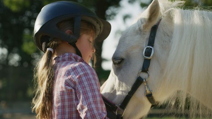 戴着骑师头盔的可爱小女孩亲吻一匹白色小马