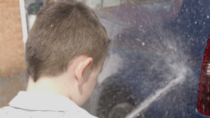 一个小男孩正在用肥皂海绵和软管冲洗车