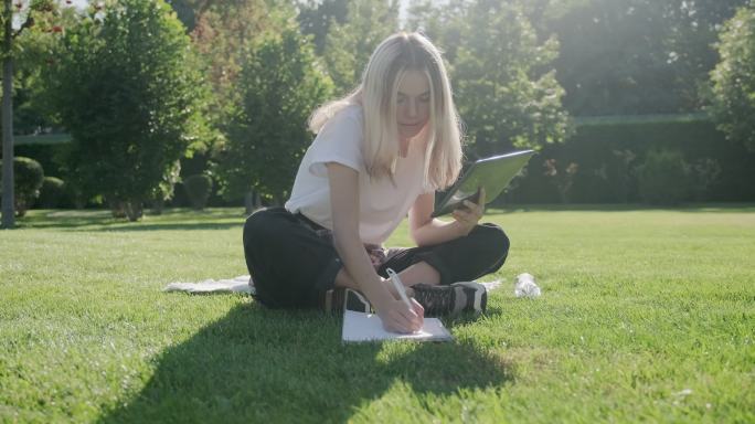 一名女生坐在公园草地上用数字平板电脑学习