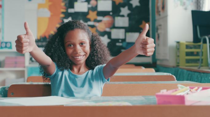 一个小女孩坐在教室的桌子旁竖起大拇指
