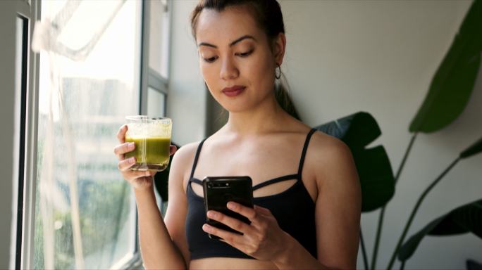 一名妇女在家使用手机时喝绿色果汁的画面