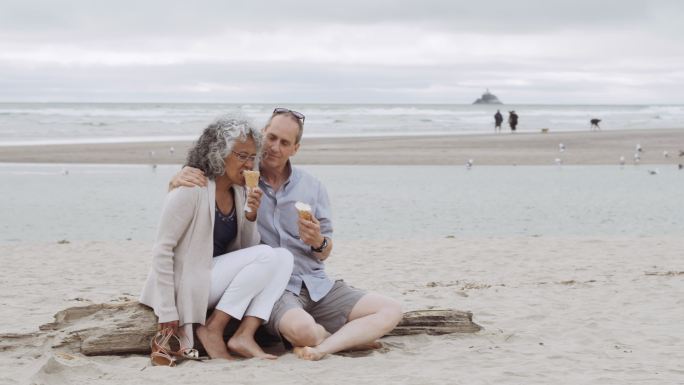 一对年长的夫妇在海滩上的圆木上吃冰淇淋