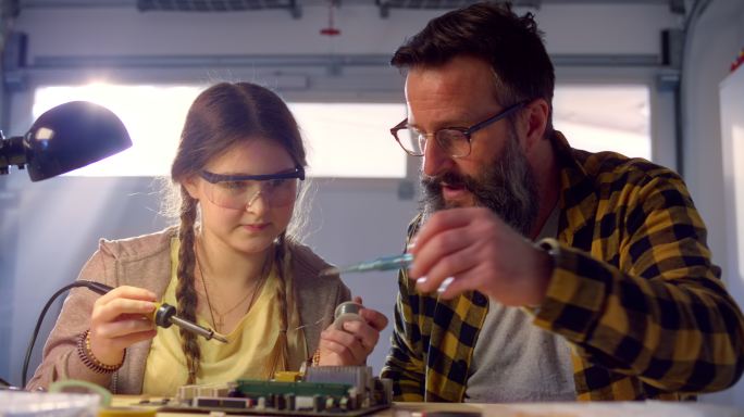 父亲将烙铁交给女儿，并指导她如何焊接