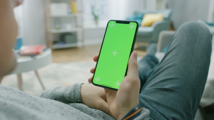 家中的男人躺在沙发上，用带有绿色模拟屏幕的智能手机