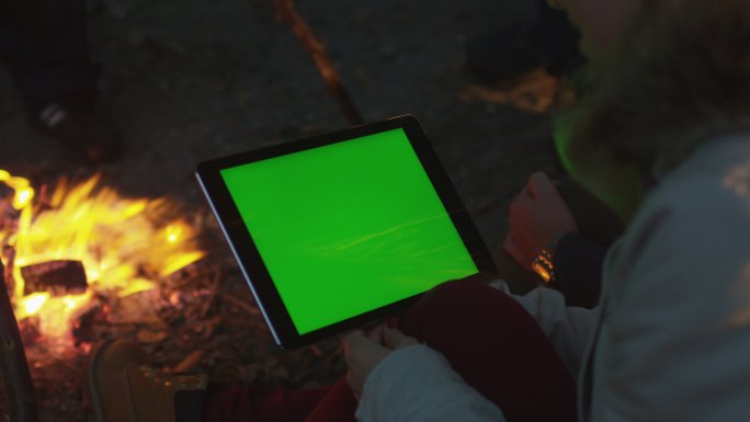 女孩在篝火旁的使用绿色屏幕电脑。