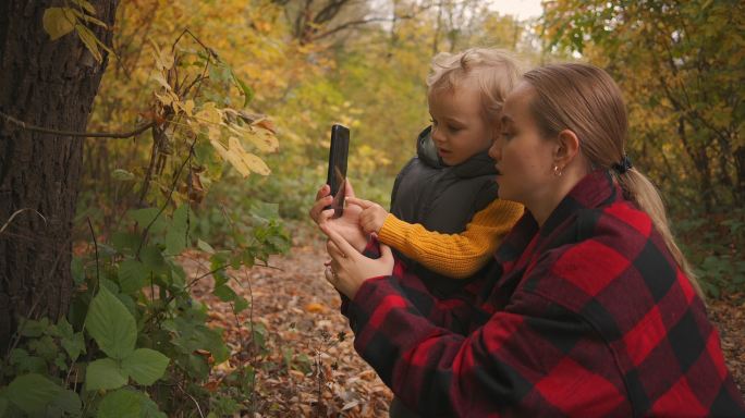 母亲在教他的小儿子在森林里用手机拍照