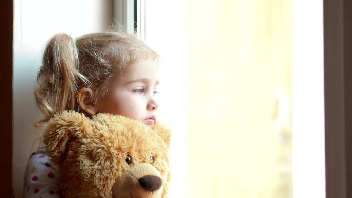 女孩怀里抱着一只泰迪熊，悲伤地凝视着窗外