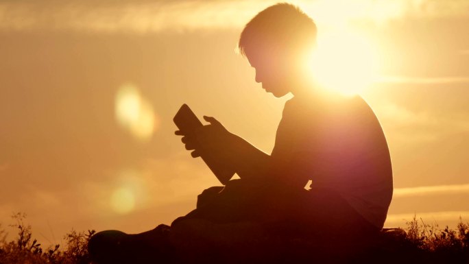 日落时坐在山上拿着平板电脑的男孩