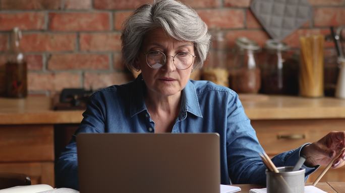 沉思的老妇人坐在室内在笔记本电脑上工作
