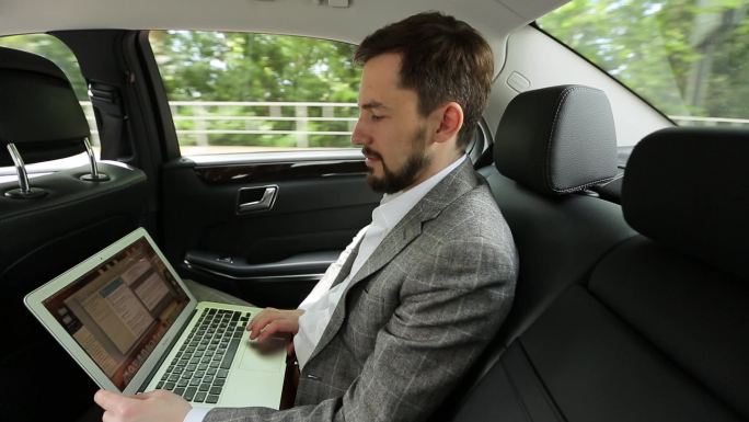 这位商人坐在汽车后座上使用笔记本电脑