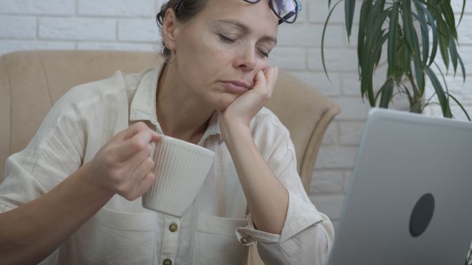 一位疲惫的女性端着一杯咖啡在电脑前睡着了