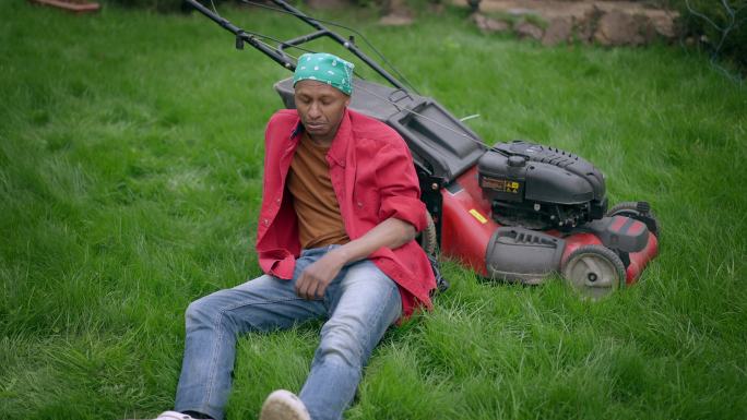 满头大汗的年轻人手拿着割草机坐在绿色的后院