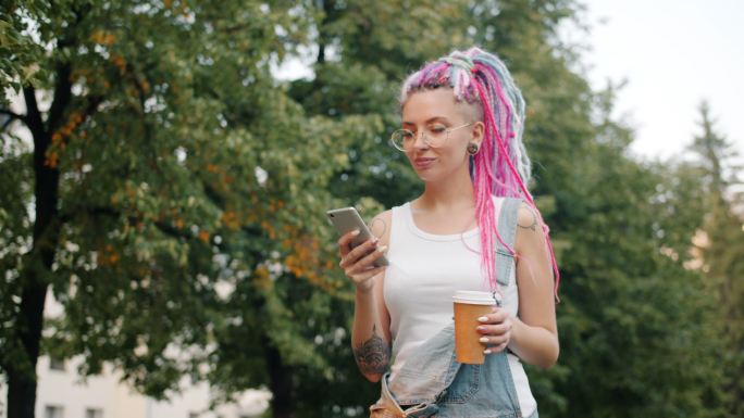 可爱的朋克女孩带着智能手机和咖啡在公园里散步