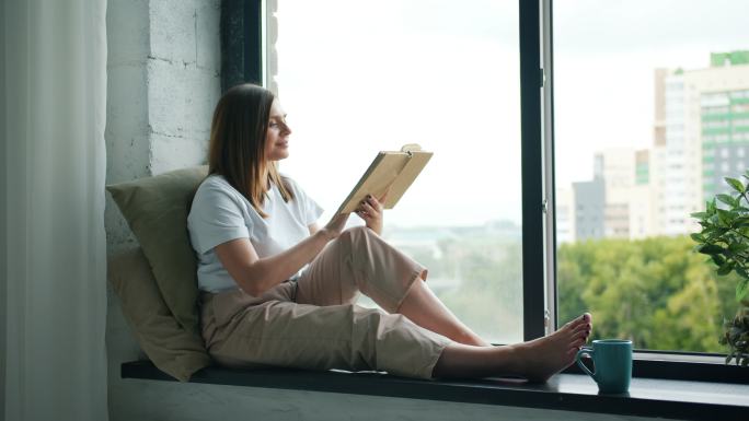 漂亮的女孩坐在家的窗台上看书翻页