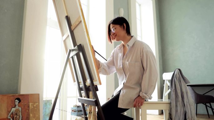 女画家坐在画架旁，用画笔在画布上画画