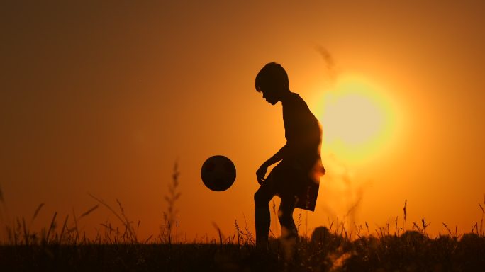 日落时一个男孩踢足球的剪影。