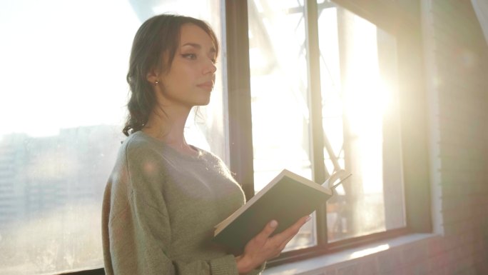 女人站在窗边，在日落的背景下看书