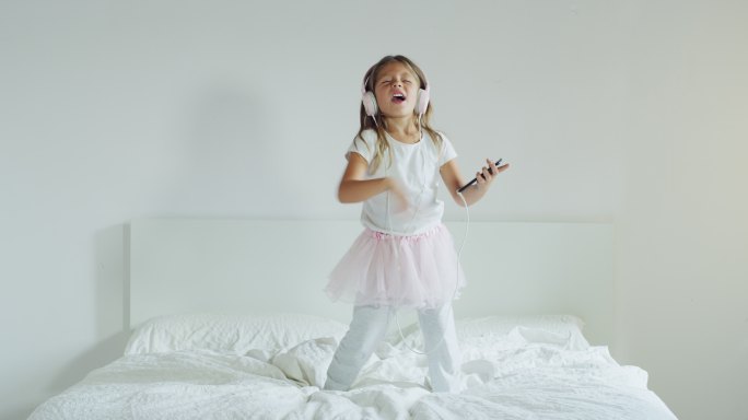 小女孩戴着耳机听音乐，在父母的床上跳舞