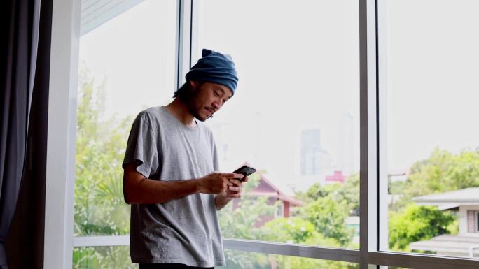 男人站在家里窗户旁，用手机输入信息。