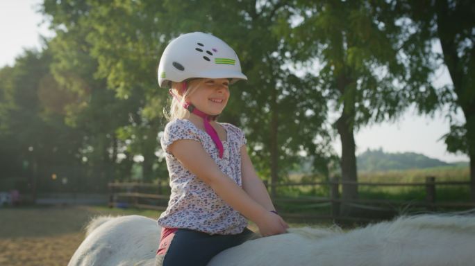 可爱小女孩骑着一匹白色小马在马厩里晒太阳