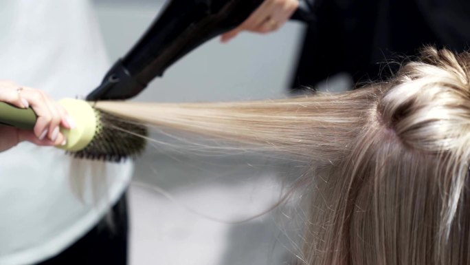 美发师用吹风机和圆梳吹干金发女人的长发