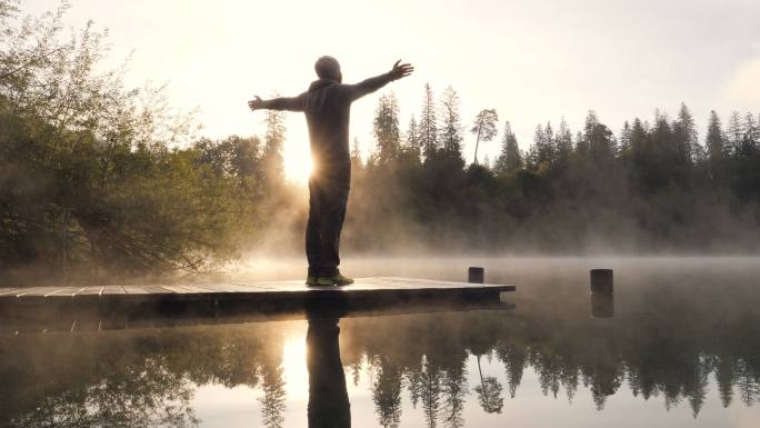 年轻人站在湖面上方的木质码头上，双臂张开