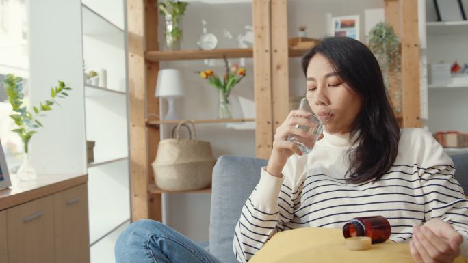 生病的年轻女子坐在家里的沙发上吃药。