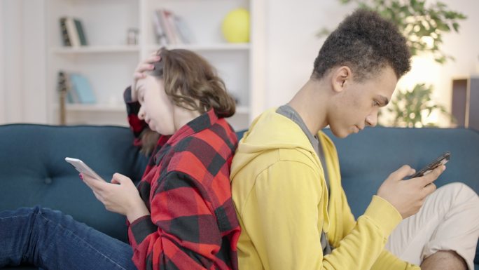 两个孩子背靠背地坐在沙发，沉迷于社交媒体
