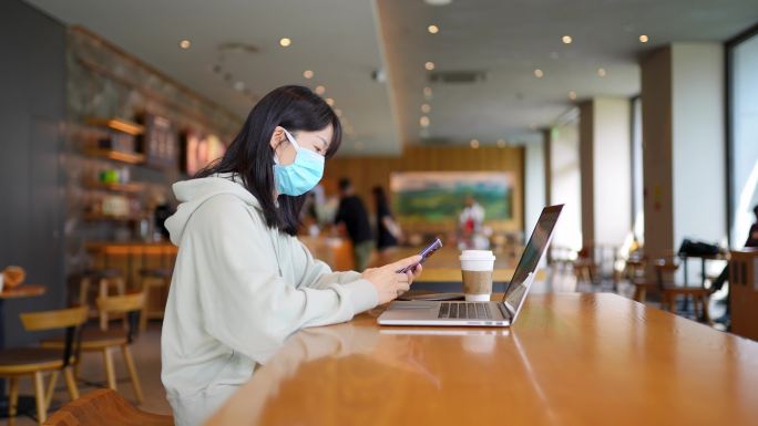 疫情带口罩年轻女子在咖啡店使用手机笔记本