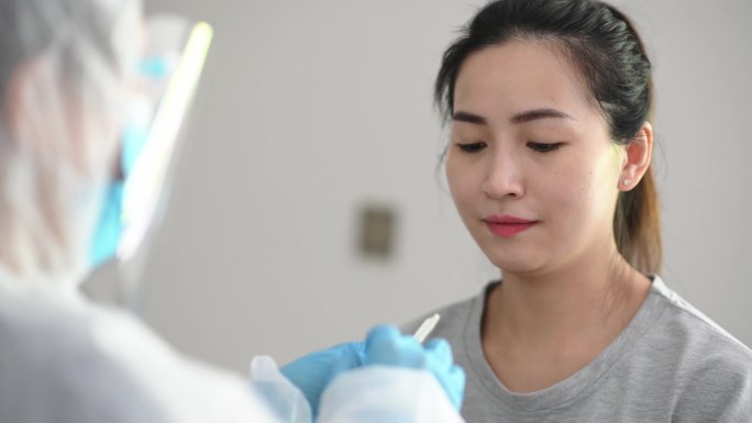携带PPE的亚裔中国女医生从患者冠状病毒检测中取鼻拭子。防护套间的医务人员正在用拭子进行冠状病毒检测
