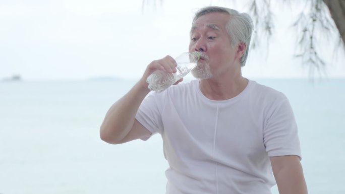 一位亚洲老人坐在海滩上放松。手里提着水瓶，带着清新的感觉喝水。照顾好自己，饮用健康水的理念